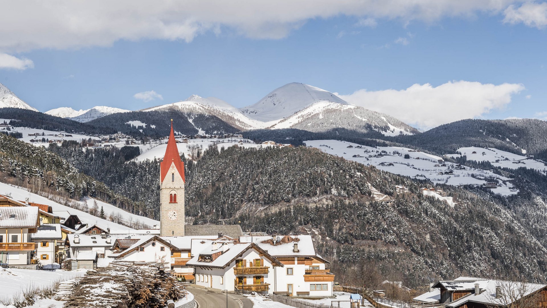 The Alpine hamlet of Spinges The picture shows a small, snow-covered village with a distinctive church and a red church tower in the foreground. In the background, forested hills and snow-covered mountains stretch beneath a blue sky with a few clouds.