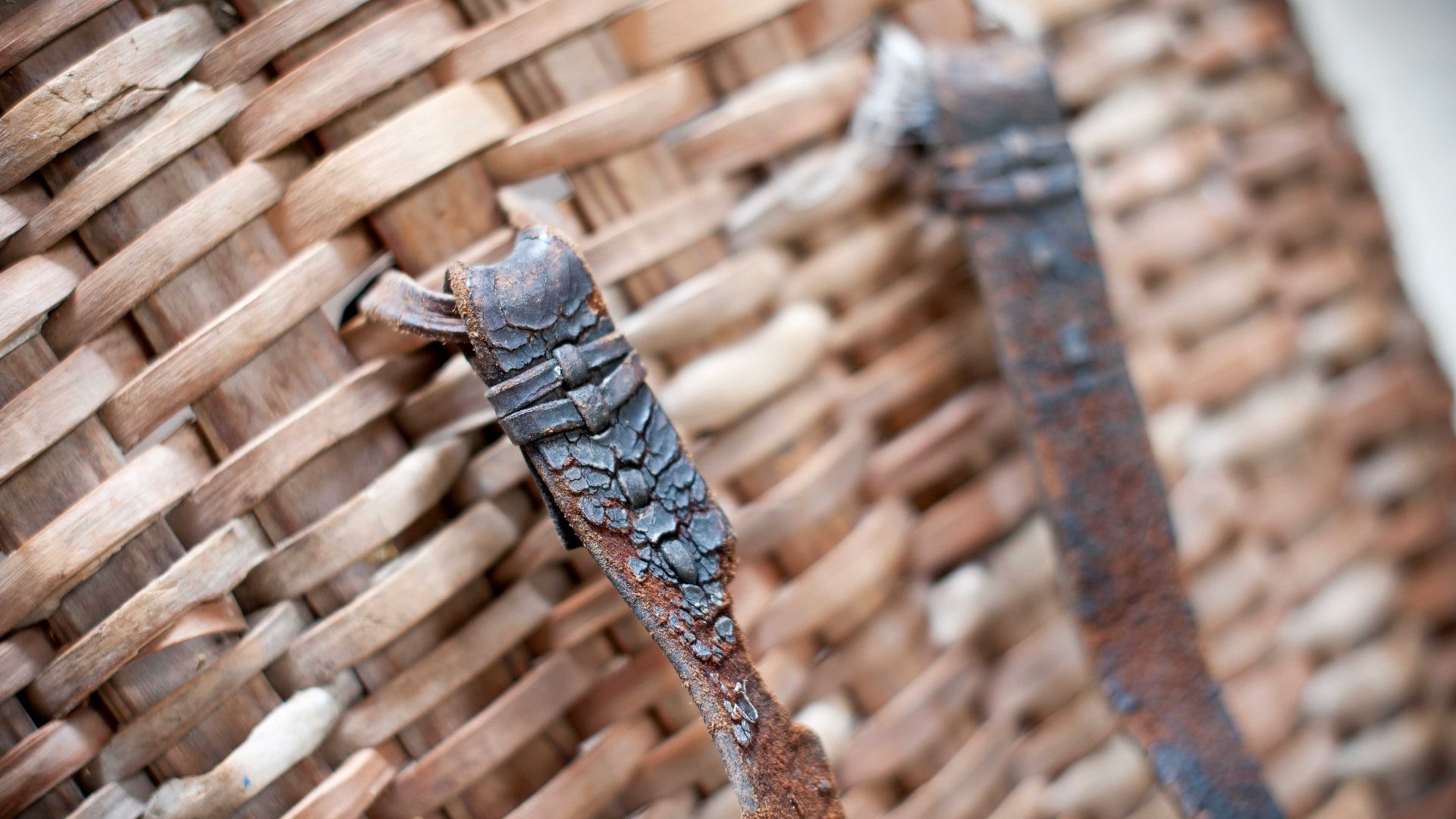 De Pfunderer Höfeweg De foto toont een close-up van een gevlochten mand gemaakt van houten strips. Twee roestige metalen banden die aan de mand zijn bevestigd, zijn op de voorgrond te zien, waarbij de structuur van de mand en de scheuren in het metaal duidelijk zichtbaar zijn.