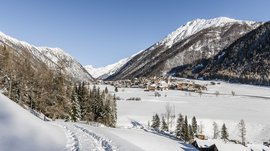 Vals: das Almendorf Das Bild zeigt eine schneebedeckte Winterlandschaft mit einem kleinen Dorf im Tal, umgeben von hohen Bergen. Ein schmaler Pfad mit Fußspuren führt durch den Schnee, während im Hintergrund klare, blaue Himmel zu sehen ist.