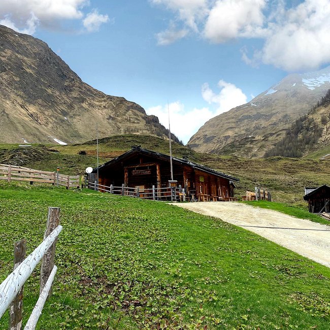 Kuttnhütte Das Bild zeigt eine rustikale Berghütte aus Holz, die auf einem sanft ansteigenden grünen Hang liegt. Die Hütte ist von einem Holzzaun umgeben, und im Hintergrund ragen hohe, felsige Berge unter einem blauen Himmel mit vereinzelten Wolken auf.