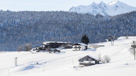 Das Bergdorf Meransen Das Bild zeigt eine winterliche Landschaft mit verschneiten Hügeln und Gebäuden, die von einem dichten, schneebedeckten Wald und hohen, schneebedeckten Bergen im Hintergrund umgeben sind. Die Häuser stehen vereinzelt auf den Hügeln und sind teils modern, teils traditionell.