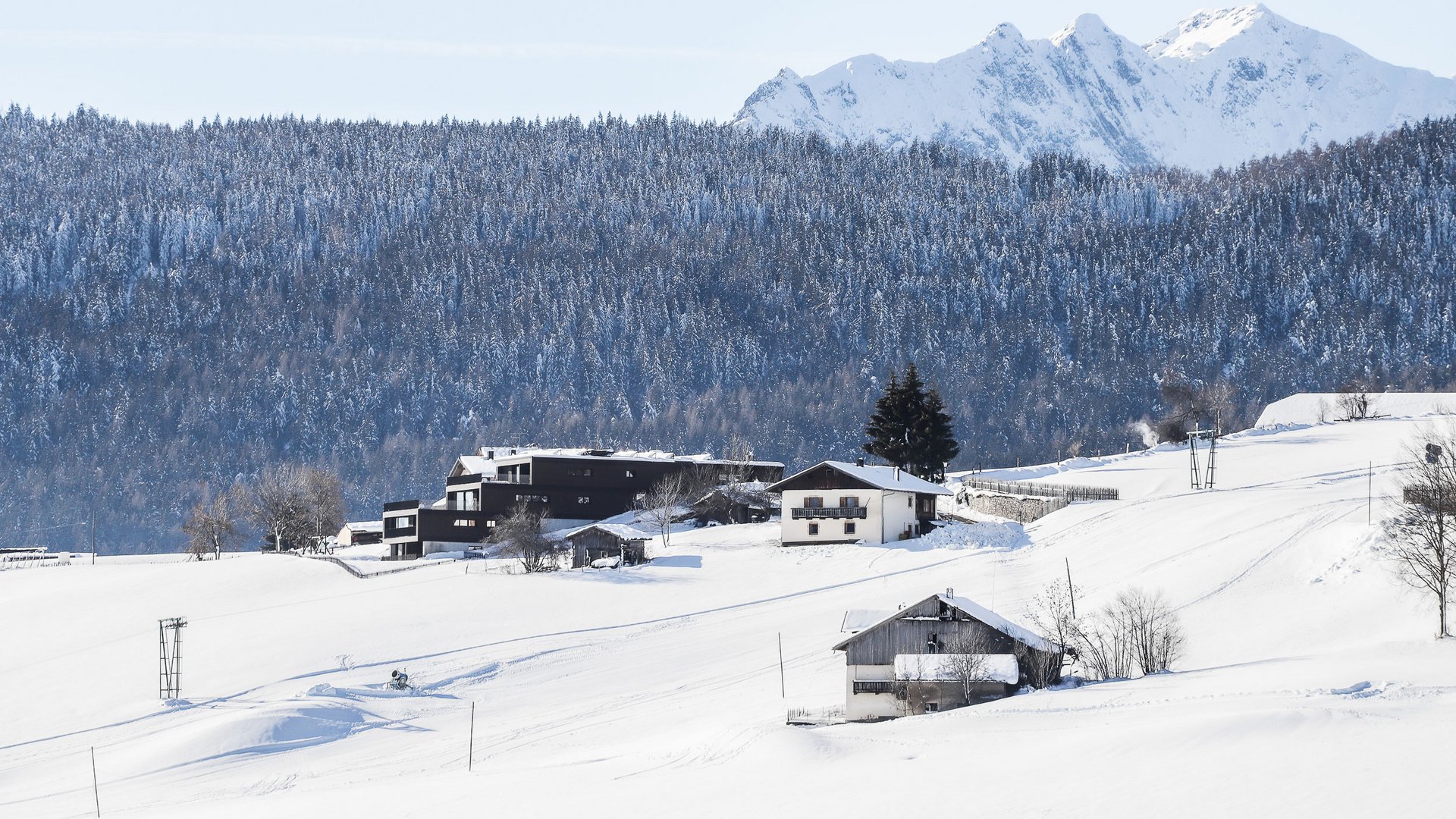 Das Bergdorf Meransen Das Bild zeigt eine winterliche Landschaft mit verschneiten Hügeln und Gebäuden, die von einem dichten, schneebedeckten Wald und hohen, schneebedeckten Bergen im Hintergrund umgeben sind. Die Häuser stehen vereinzelt auf den Hügeln und sind teils modern, teils traditionell.