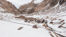 Fane Alm – nejkrásnější horská vesnice Jižního Tyrolska Obrázek ukazuje malé sídliště tradičních dřevěných domů, které leží v údolí, obklopené zasněženými horami. Sníh začíná tát a objevují se hnědé horské plochy, zatímco oblast obklopuje plot.