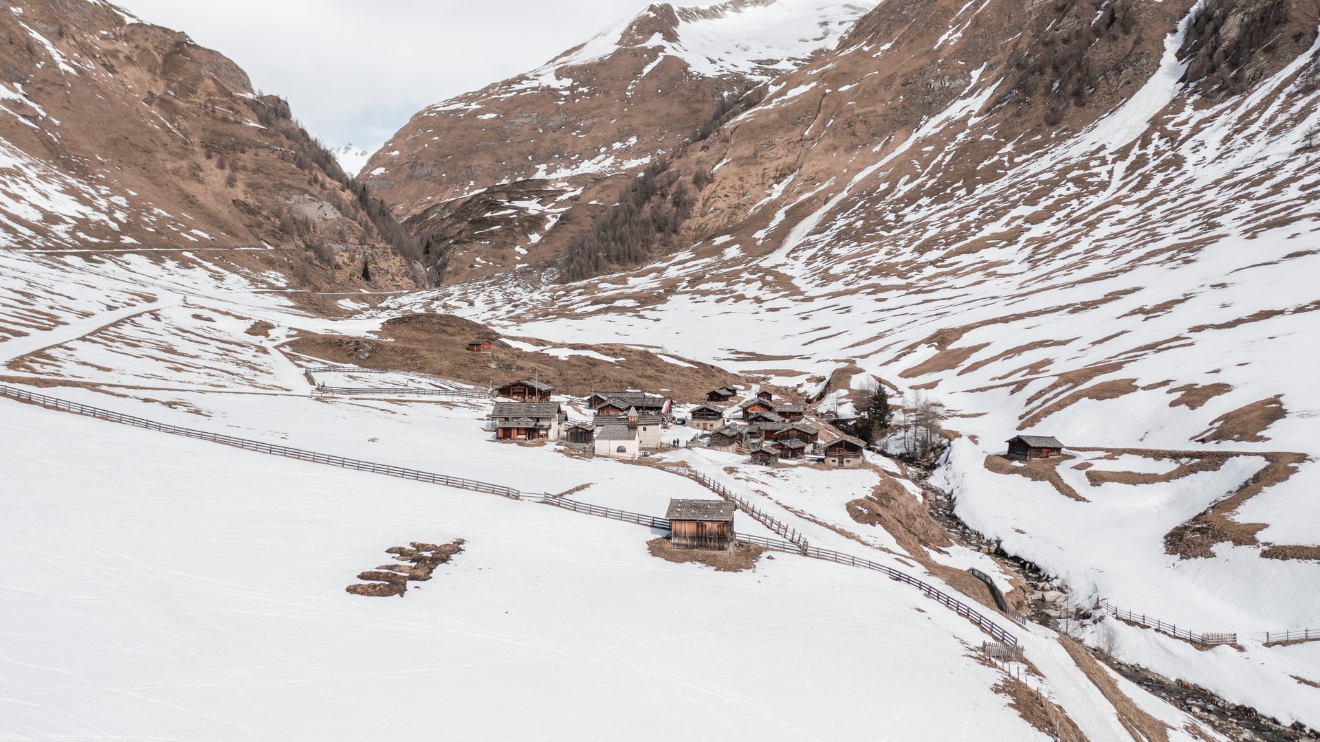 Fane Alm – nejkrásnější horská vesnice Jižního Tyrolska Obrázek ukazuje malé sídliště tradičních dřevěných domů, které leží v údolí, obklopené zasněženými horami. Sníh začíná tát a objevují se hnědé horské plochy, zatímco oblast obklopuje plot.