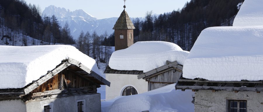 Find your accommodation in South Tyrol The picture shows a snowy mountain hut settlement with deep snow on the roofs of the buildings. In the background, there is a small church tower with a pointed roof, surrounded by snow-covered trees and mountains.