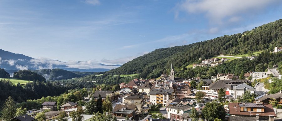 Find your accommodation in South Tyrol The picture shows a small, picturesque town in a green valley, surrounded by wooded hills and mountains. In the center of the picture stands a church tower with a spire, and the buildings are surrounded by lush vegetation and some scattered clouds.