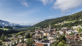 Mühlbach in the Pustertal valley The picture shows a small, picturesque town in a green valley, surrounded by wooded hills and mountains. In the center of the picture stands a church tower with a spire, and the buildings are surrounded by lush vegetation and some scattered clouds.