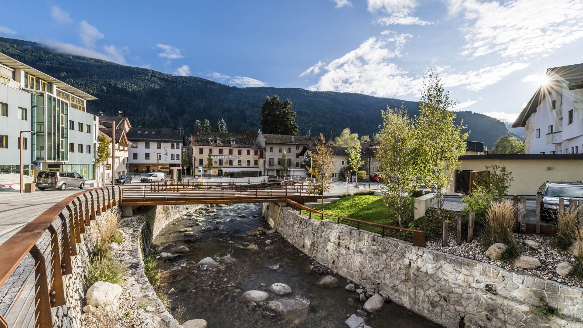 Vintl in het Pustertal De foto toont een kleine brug die over een heldere beek in een dorp loopt. Op de achtergrond zijn huizen en groene heuvels of bergen te zien onder een blauwe lucht met enkele wolken.