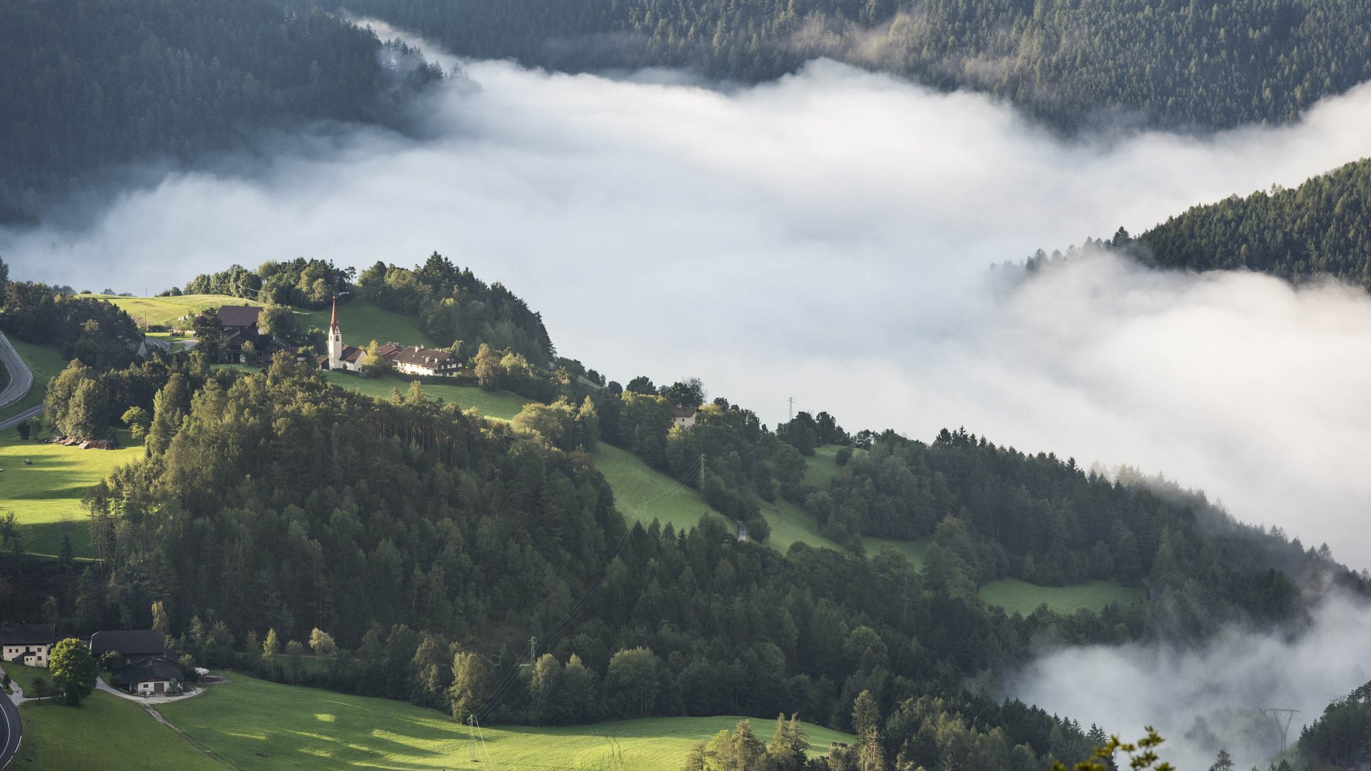 Rodeneck – for a wonderful walking holiday in Italy The picture shows a hilly landscape pierced by dense white fog. On one of the hills stands a small church, surrounded by green meadows and forests that rise out of the fog.