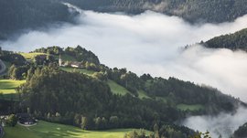 Rodeneck – for a wonderful walking holiday in Italy The picture shows a hilly landscape pierced by dense white fog. On one of the hills stands a small church, surrounded by green meadows and forests that rise out of the fog.