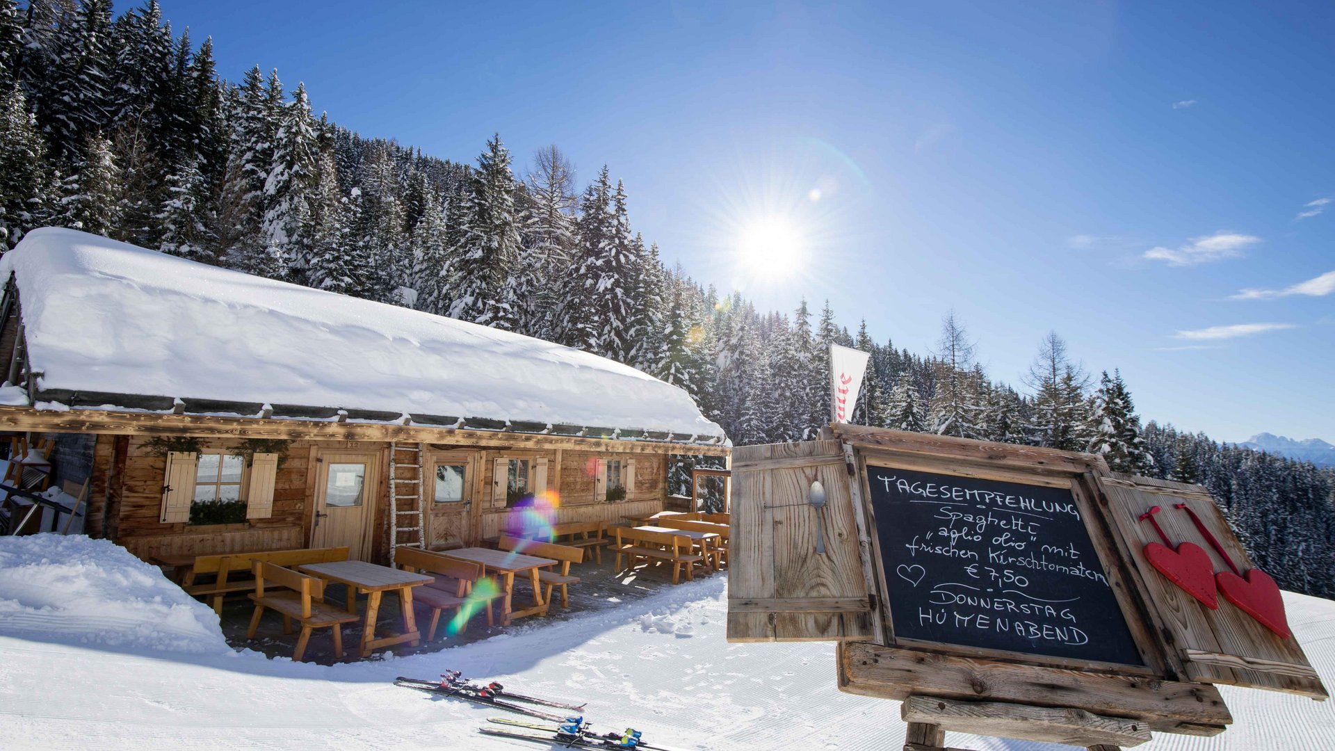 Gitschberg Jochtal holiday area – mountain huts and shelters The picture shows a snow-covered mountain hut with wooden benches and tables in front of the hut; next to it is a board displaying the daily special, while in the background snow-covered coniferous trees and a bright blue sky can be seen.