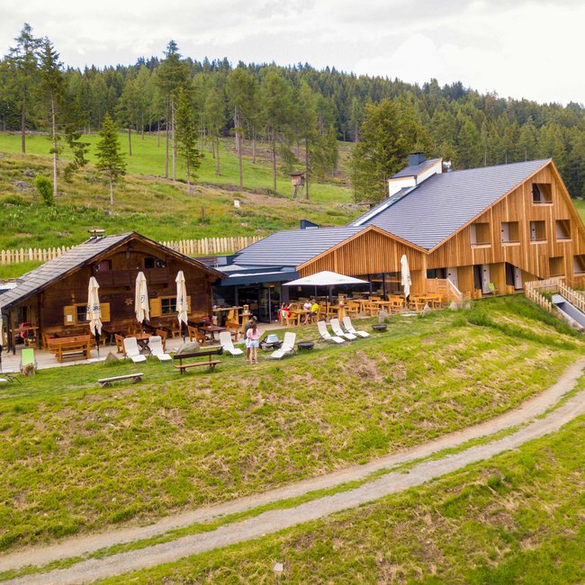 Oberhauserhütte De afbeelding toont een moderne houten berghut, omgeven door groene weiden en bossen. Voor de hut staan talrijke ligstoelen en tafels met parasols, die uitnodigen om te ontspannen in de natuur.