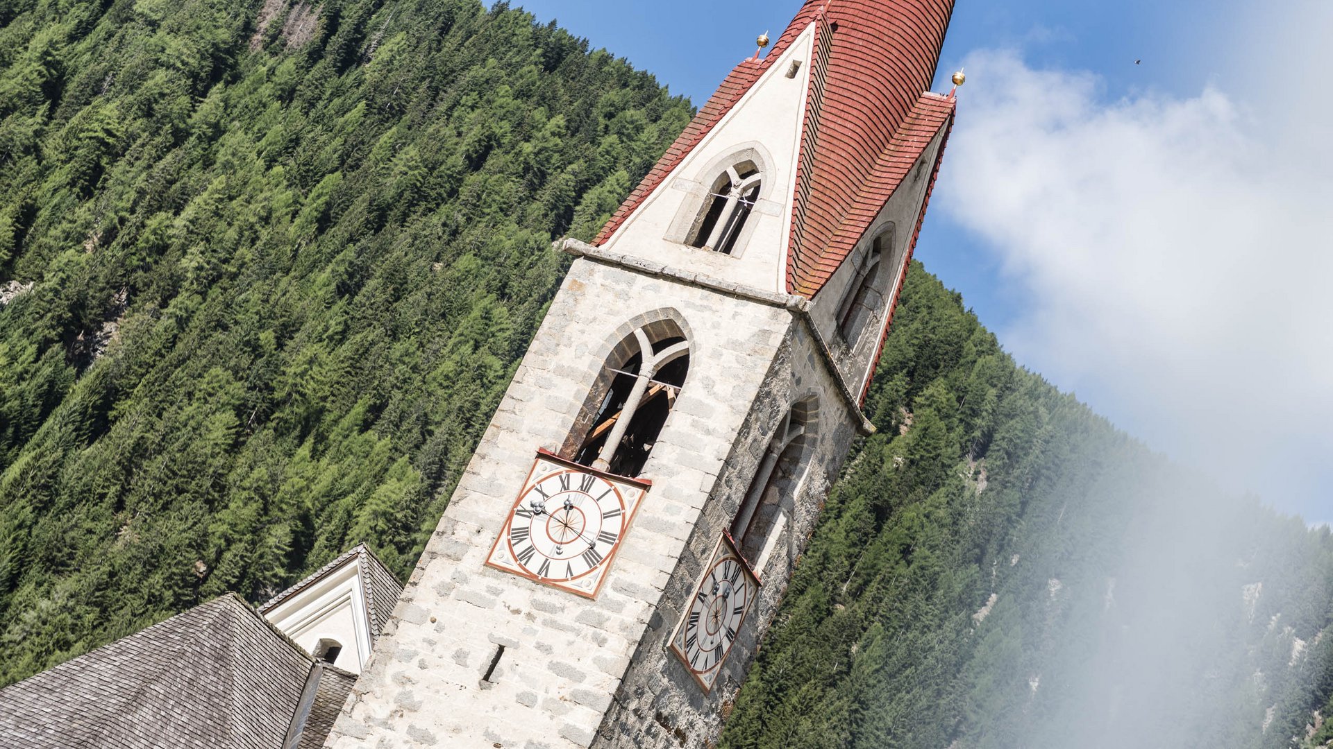 Vals: das Almendorf Das Bild zeigt einen Kirchturm mit einer roten Spitze und zwei großen Uhren mit römischen Ziffern. Im Hintergrund sind dichte grüne Nadelwälder zu sehen, und der Himmel ist teils bewölkt.