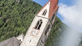 Vals: das Almendorf Das Bild zeigt einen Kirchturm mit einer roten Spitze und zwei großen Uhren mit römischen Ziffern. Im Hintergrund sind dichte grüne Nadelwälder zu sehen, und der Himmel ist teils bewölkt.