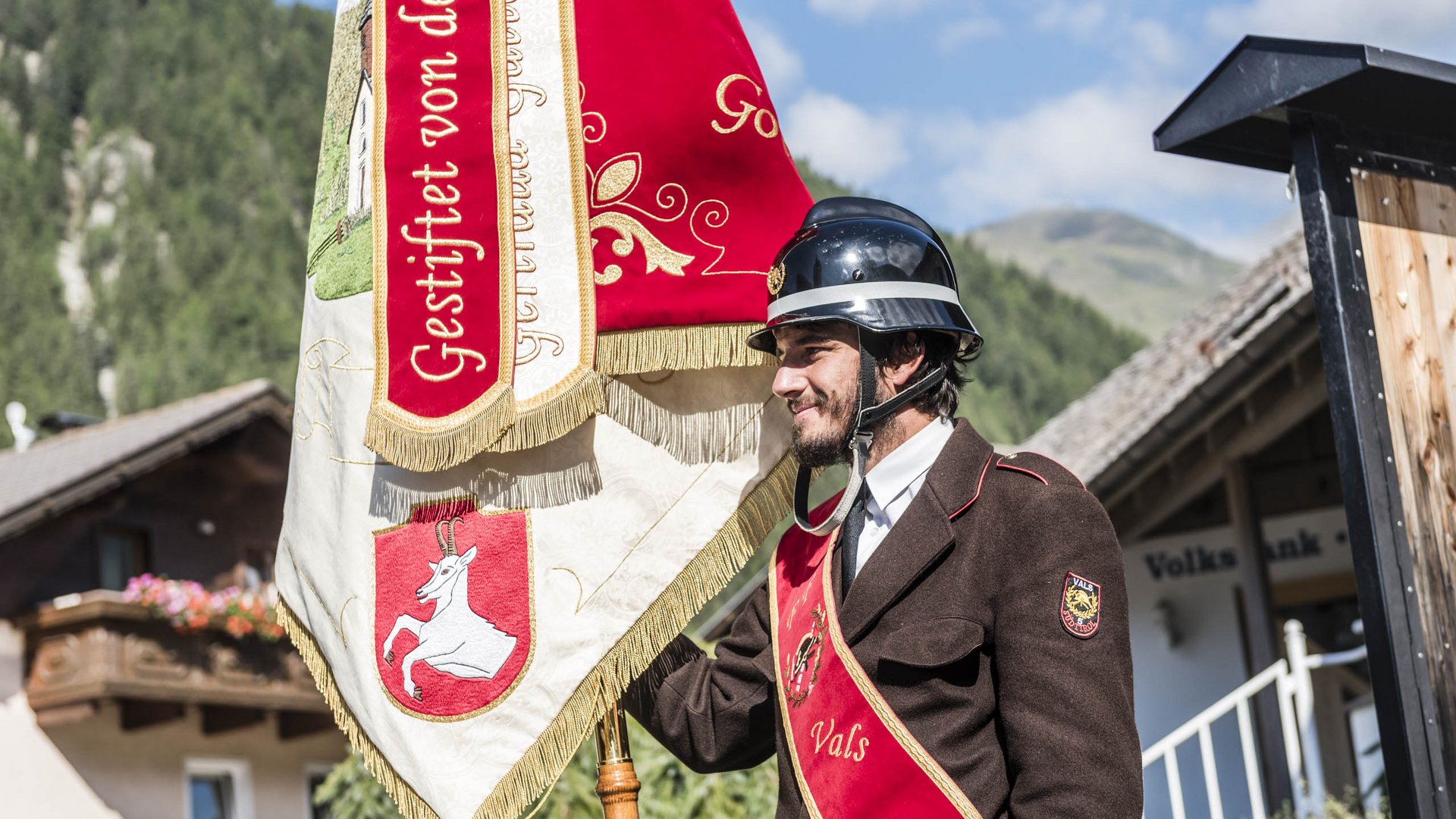 Vals: das Almendorf Das Bild zeigt einen Mann in einer braunen Uniform mit roter Schärpe, auf der "Vals" steht. Er trägt einen schwarzen Helm und hält eine rot-weiße Fahne mit goldenen Verzierungen und einer weißen Ziege darauf. Im Hintergrund sind ein Haus und Berge zu sehen.