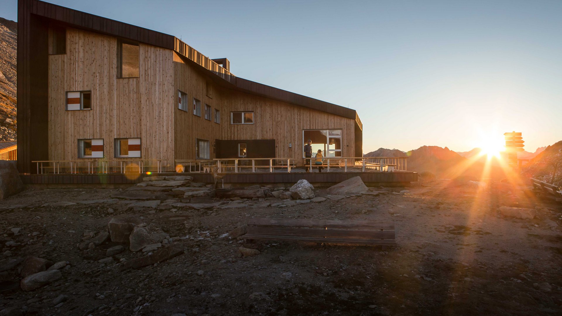 The Pfunders High Mountain trail The picture shows a modern wooden mountain hut in the mountains at sunset. The hut has large windows and a terrace, while the sun sets behind the mountains, bathing the surroundings in warm light.