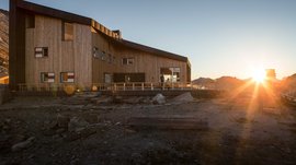 The Pfunders High Mountain trail The picture shows a modern wooden mountain hut in the mountains at sunset. The hut has large windows and a terrace, while the sun sets behind the mountains, bathing the surroundings in warm light.