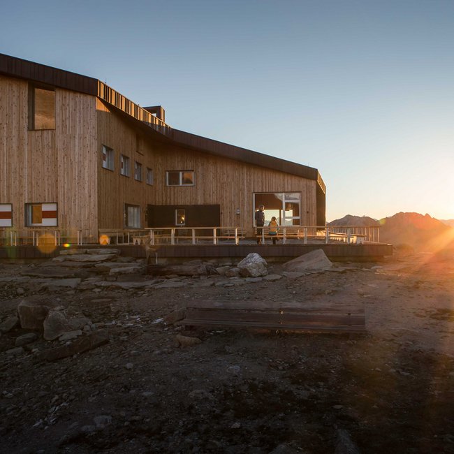 Edelrauthütte The picture shows a modern wooden mountain hut in the mountains at sunset. The hut has large windows and a terrace, while the sun sets behind the mountains, bathing the surroundings in warm light.