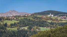 Das Bergdorf Meransen Das Bild zeigt eine malerische Landschaft mit einem Dorf, das sich auf grünen Hügeln ausbreitet. Im Vordergrund sind dichte Wälder zu sehen, während im Hintergrund hohe Berge unter einem klaren, blauen Himmel aufragen. Die Häuser und eine Kirche des Dorfes sind inmitten der grünen Hügel verstreut.