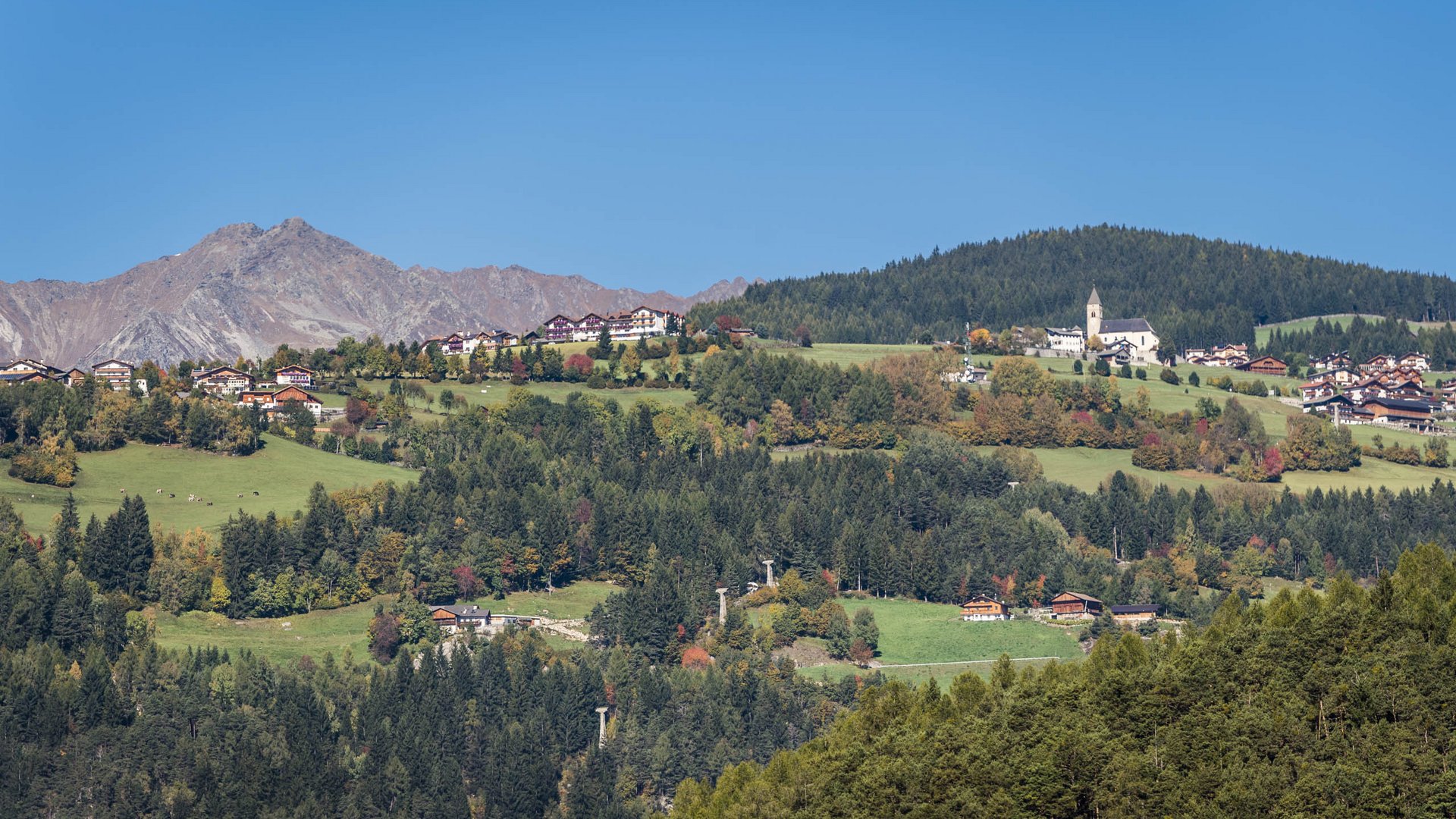 Das Bergdorf Meransen Das Bild zeigt eine malerische Landschaft mit einem Dorf, das sich auf grünen Hügeln ausbreitet. Im Vordergrund sind dichte Wälder zu sehen, während im Hintergrund hohe Berge unter einem klaren, blauen Himmel aufragen. Die Häuser und eine Kirche des Dorfes sind inmitten der grünen Hügel verstreut.