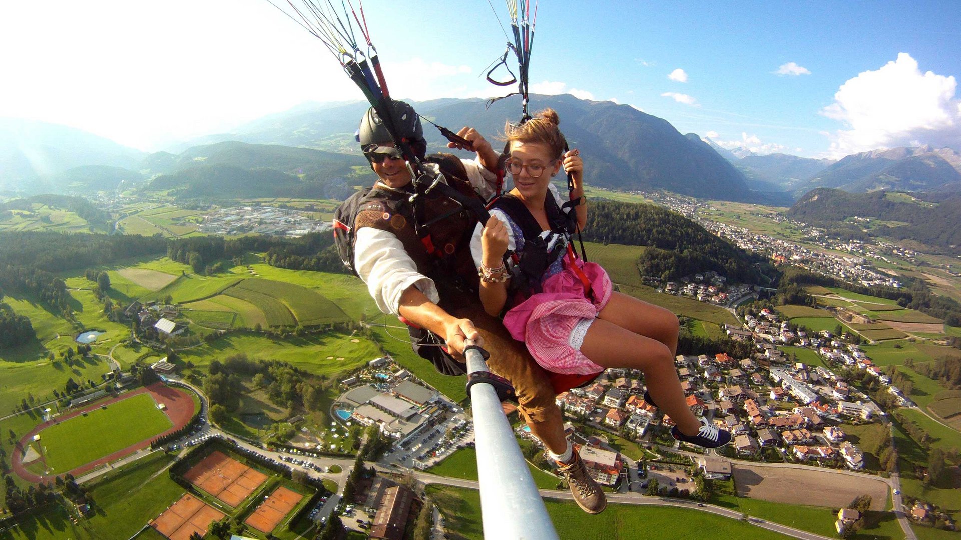 Paragliden in Zuid-Tirol De afbeelding toont twee personen die samen in de lucht aan het paragliden zijn. Onder hen strekt zich een groen landschap uit met velden, een sportveld en een klein stadje. De personen lijken plezier te hebben, en de piloot houdt een camera of een selfie-stick vast om het moment vast te leggen.
