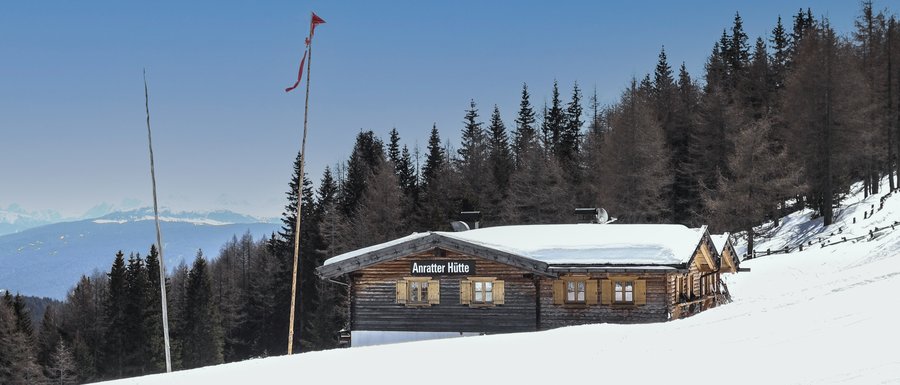Find your accommodation in South Tyrol The picture shows the "Anratter Hütte," a wooden hut on a snow-covered slope, surrounded by coniferous trees. In the background, snow-covered mountain peaks are visible under a blue sky. A red flag flies on one of the flagpoles in front of the hut.