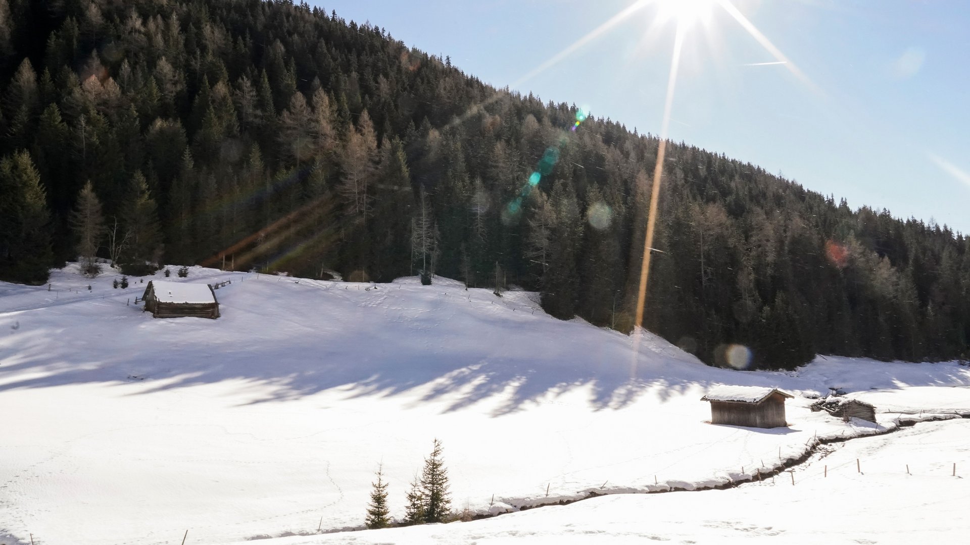 Het Altfassdal bij Meransen De afbeelding toont een zonnig winterlandschap met twee kleine, met sneeuw bedekte houten hutten in een dal. Op de achtergrond rijst een dicht, met sneeuw bedekt bos op, en de stralende zon staat hoog aan de heldere blauwe hemel en werpt lange schaduwen op de sneeuw.