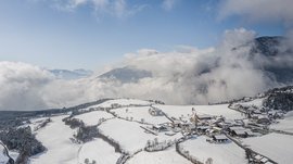 The Alpine hamlet of Spinges The picture shows a snowy mountain landscape with a small village in the foreground. The sky is clear, and clouds are moving over the mountains in the background, while the houses and fields of the village are covered with snow.