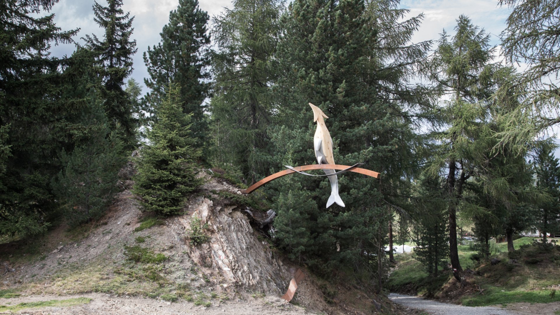 The Creation trail in Rodeneck The picture shows an art installation in a forest area. The sculpture consists of an abstracted fish and a bird balancing on a curved metal arch.