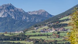 The Alpine hamlet of Spinges The picture shows a picturesque landscape with green meadows, scattered houses, and a small church with a red tower. In the background, wooded mountain slopes rise, topped by steep, rocky peaks.