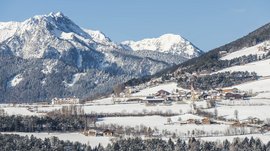 The Alpine hamlet of Spinges The picture shows a winter landscape with a snow-covered village in the foreground and snow-covered mountains in the background. The village is surrounded by forests and meadows, and a church with a red tower stands out.