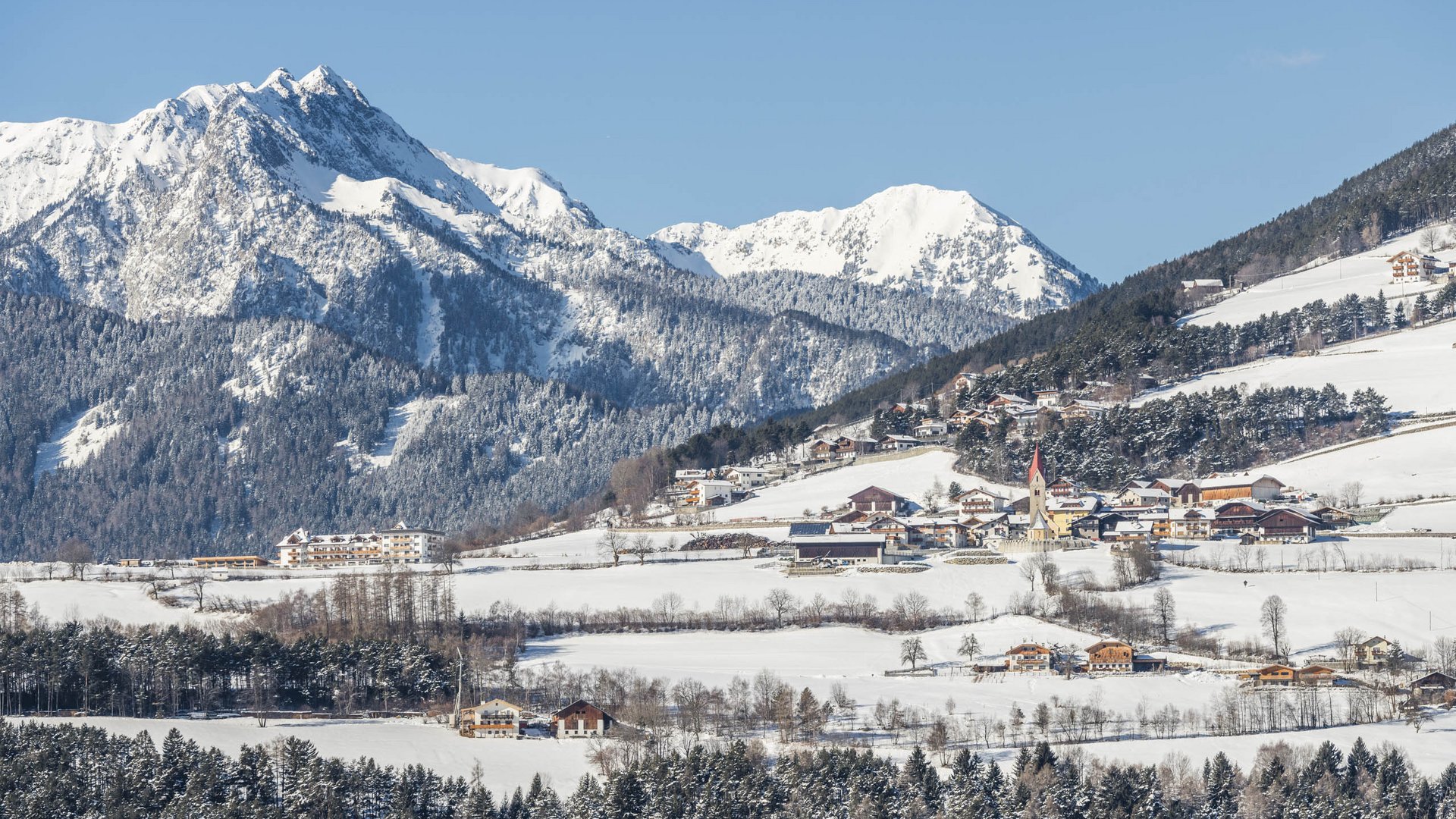 The Alpine hamlet of Spinges The picture shows a winter landscape with a snow-covered village in the foreground and snow-covered mountains in the background. The village is surrounded by forests and meadows, and a church with a red tower stands out.