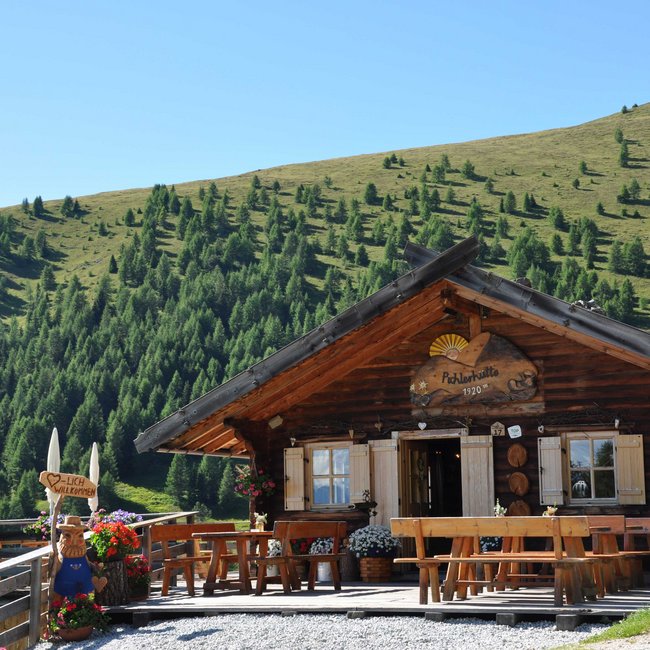 Pichlerhütte Obrázek ukazuje útulnou horskou chatu ze dřeva, obklopenou zelenými loukami a mnoha smrky. Před chatou stojí dřevěné lavice a květináče s barevnými květinami. Dřevěná cedule s nápisem "Srdečně vítáme" vítá návštěvníky.