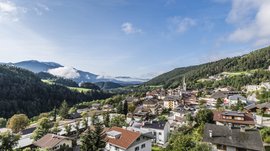 Mühlbach in the Pustertal valley The picture shows a picturesque town surrounded by green hills and mountains. In the foreground, houses with red roofs and lush greenery can be seen, while in the background fog-covered mountains and a clear blue sky are visible.