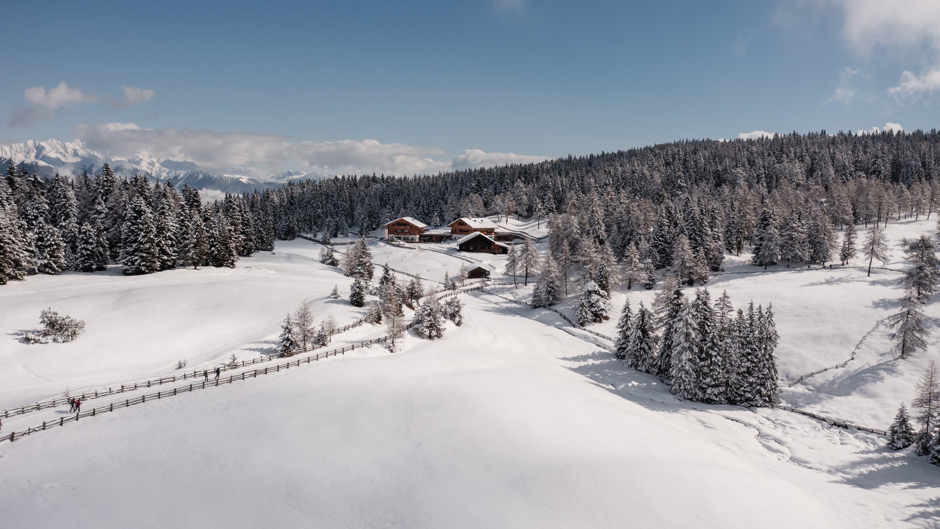 De Rodenecker-Lüsner Alm De foto toont een besneeuwd winterlandschap met meerdere hutten die omringd zijn door een dikke sneeuwlaag. Op de voorgrond loopt een besneeuwd pad langs een houten hek, en op de achtergrond strekt zich een dicht bos uit met met sneeuw bedekte bomen. De lucht is helder en blauw, wat een rustige en vredige sfeer uitstraalt.
