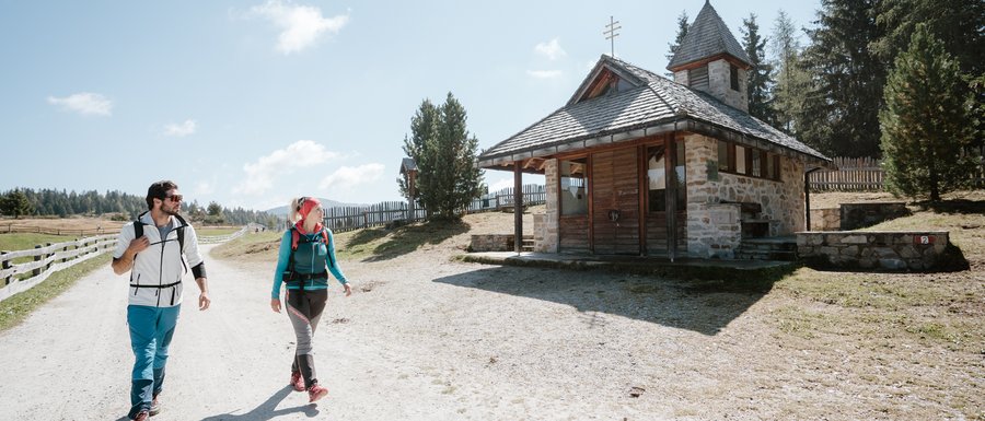 Find your accommodation in South Tyrol The picture shows two hikers, a man and a woman, walking along a wide, sandy path. To their right stands a small stone chapel with a wooden roof. The sky is clear and blue, and in the background, gentle hills and trees can be seen.