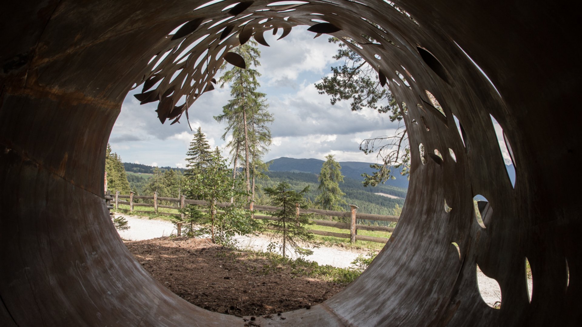 The Creation trail in Rodeneck The picture shows the view through a large, rust-colored metal pipe, artistically pierced with leaf patterns. Outside there are trees, a wooden fence, and hills in the distance under a cloudy sky.
