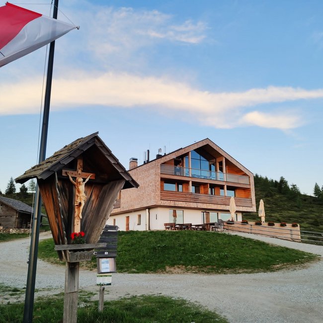 Starkenfeldhütte De afbeelding toont een modern, drie verdiepingen tellend berghuis met grote ramen en houten bekleding, dat op een heuvel staat. Op de voorgrond wappert een rood-wit vlag naast een houten kruisbeeld, en de lucht is licht bewolkt met een zachte avondlicht.