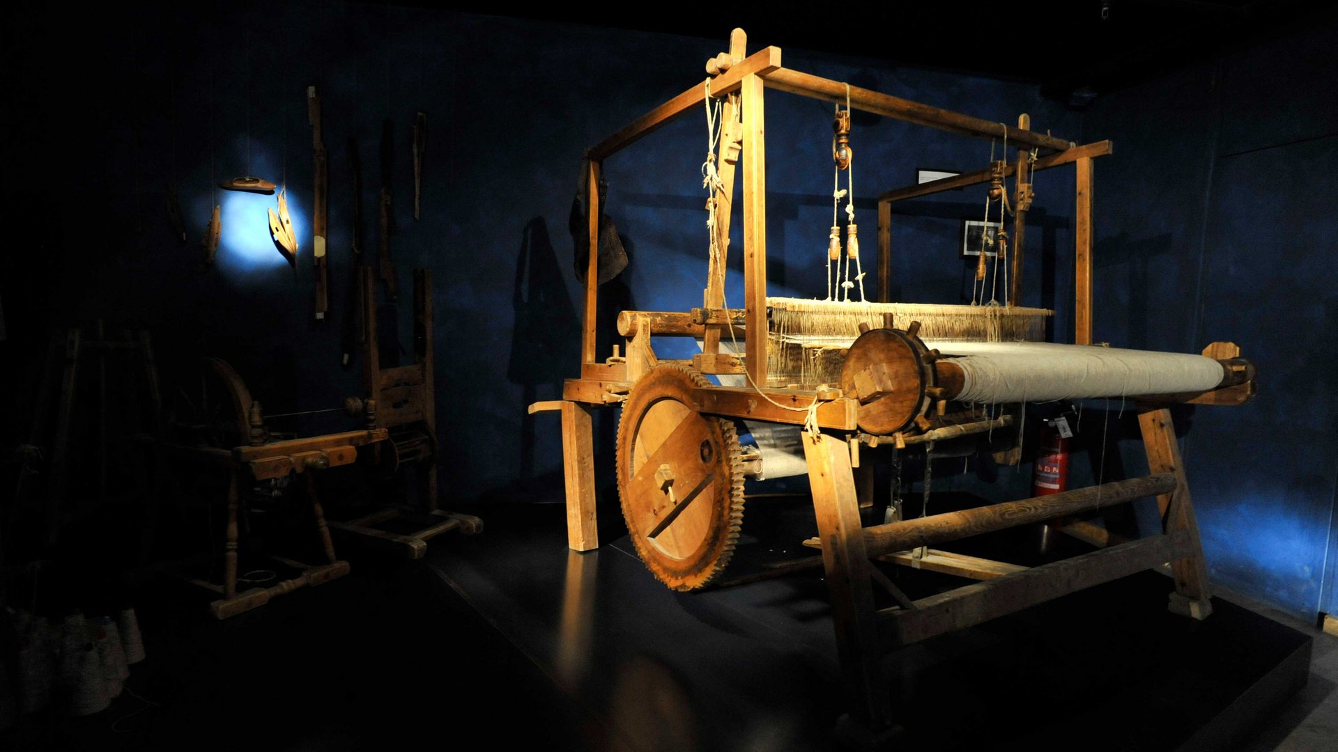Zážitkový svět lodenu ve Vintlu/Pustertal Obrázek ukazuje velký dřevěný stav v tmavé místnosti, kterou osvětluje světelný paprsek. V pozadí jsou na zdi vidět další historické nástroje.
