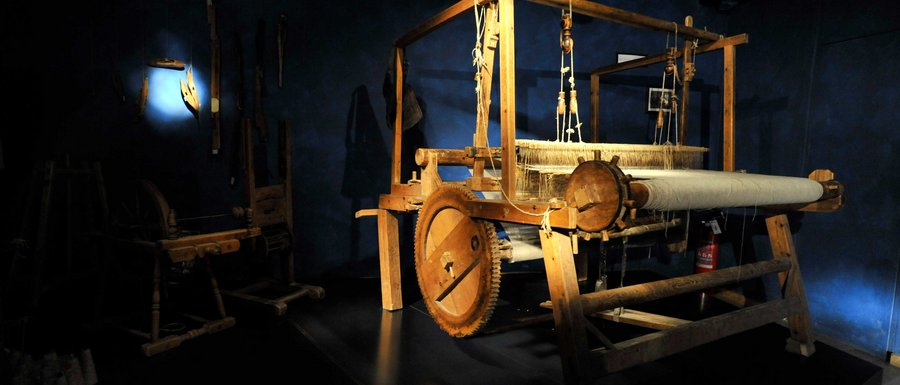 Find your accommodation in South Tyrol The image shows a large wooden loom in a dark room, illuminated by a beam of light. In the background, other historical tools can be seen on the wall.