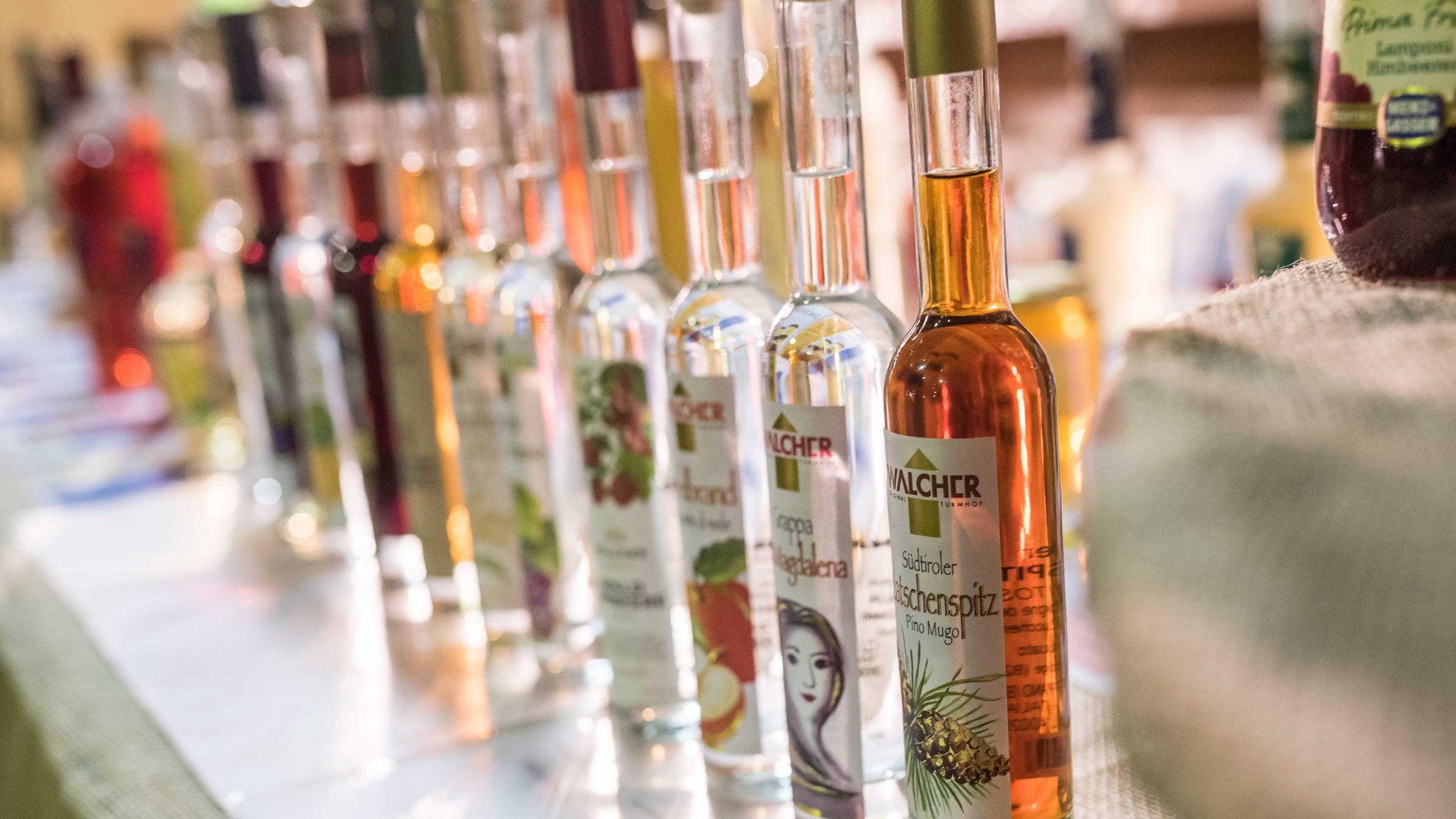 Nakupování v Mühlbachu/Itálie Obrázek ukazuje řadu lahví s různými lihovinami, které jsou těsně vedle sebe postavené. Každá láhev má etiketu s různými motivy a texty, které naznačují různé příchutě nebo složky.