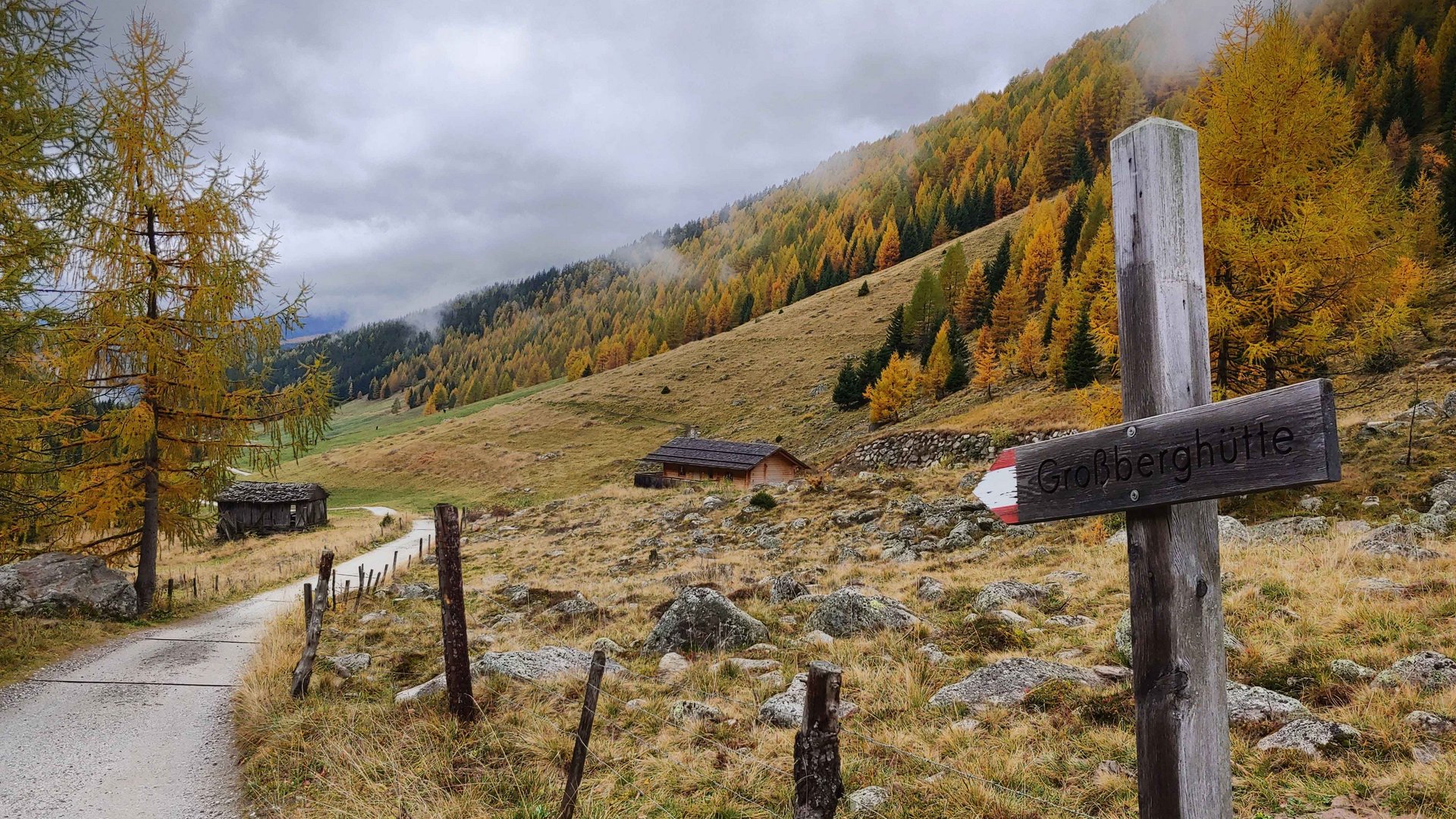Het Altfassdal bij Meransen De afbeelding toont een wandelpad dat door een herfstachtig landschap met kleurrijke, deels gele en oranje bomen loopt. De lucht is bewolkt en aan de kant van het pad staat een houten wegwijzer met de tekst "Großberghütte." Op de achtergrond zijn een paar hutten en een beboste heuvel te zien.