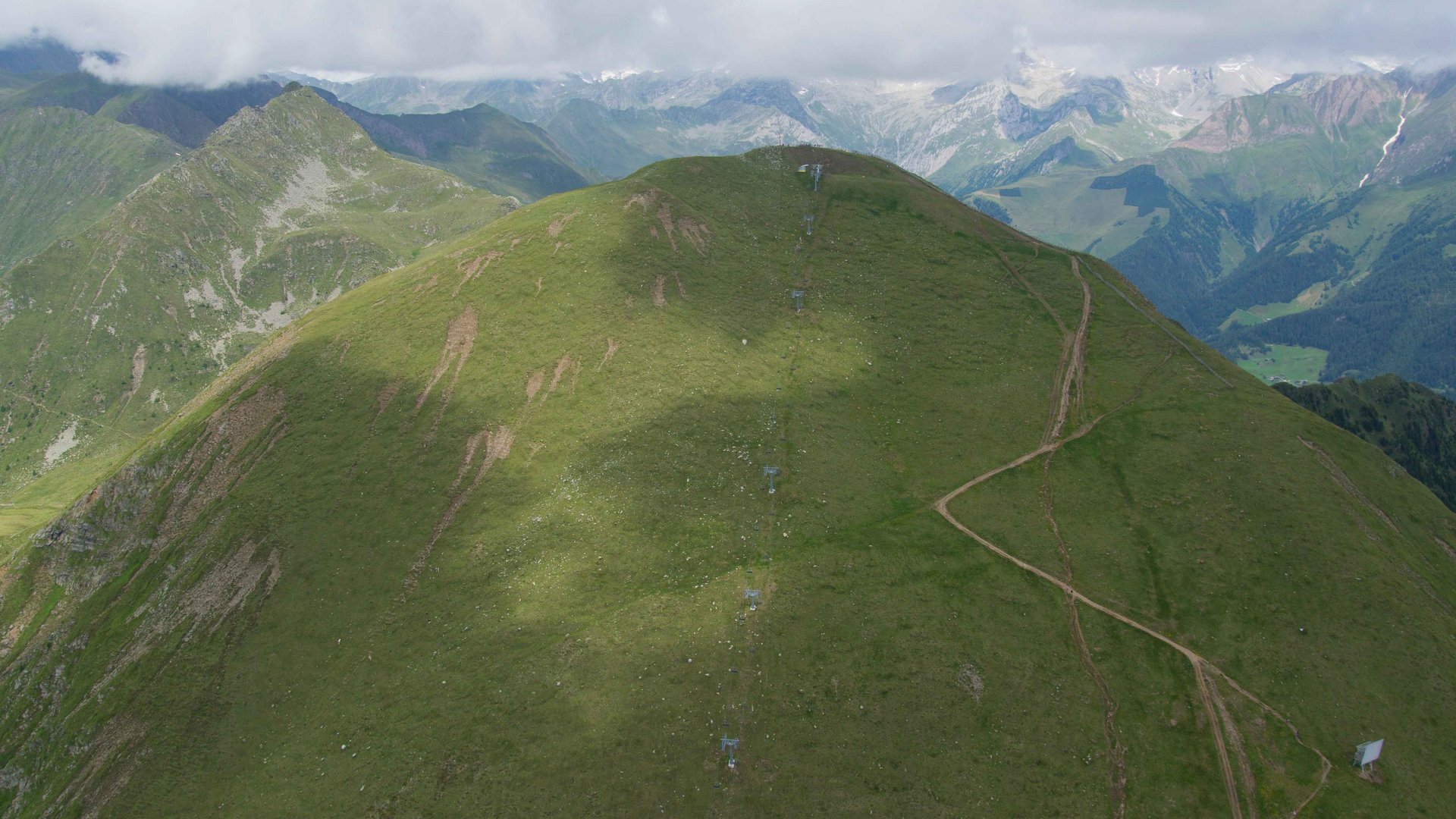 The Gitschberg panoramic platform in Meransen, Italy The picture shows a green, grass-covered mountain ridge with a narrow path running up the slope. In the background, additional mountains and valleys are visible, partially covered by clouds.