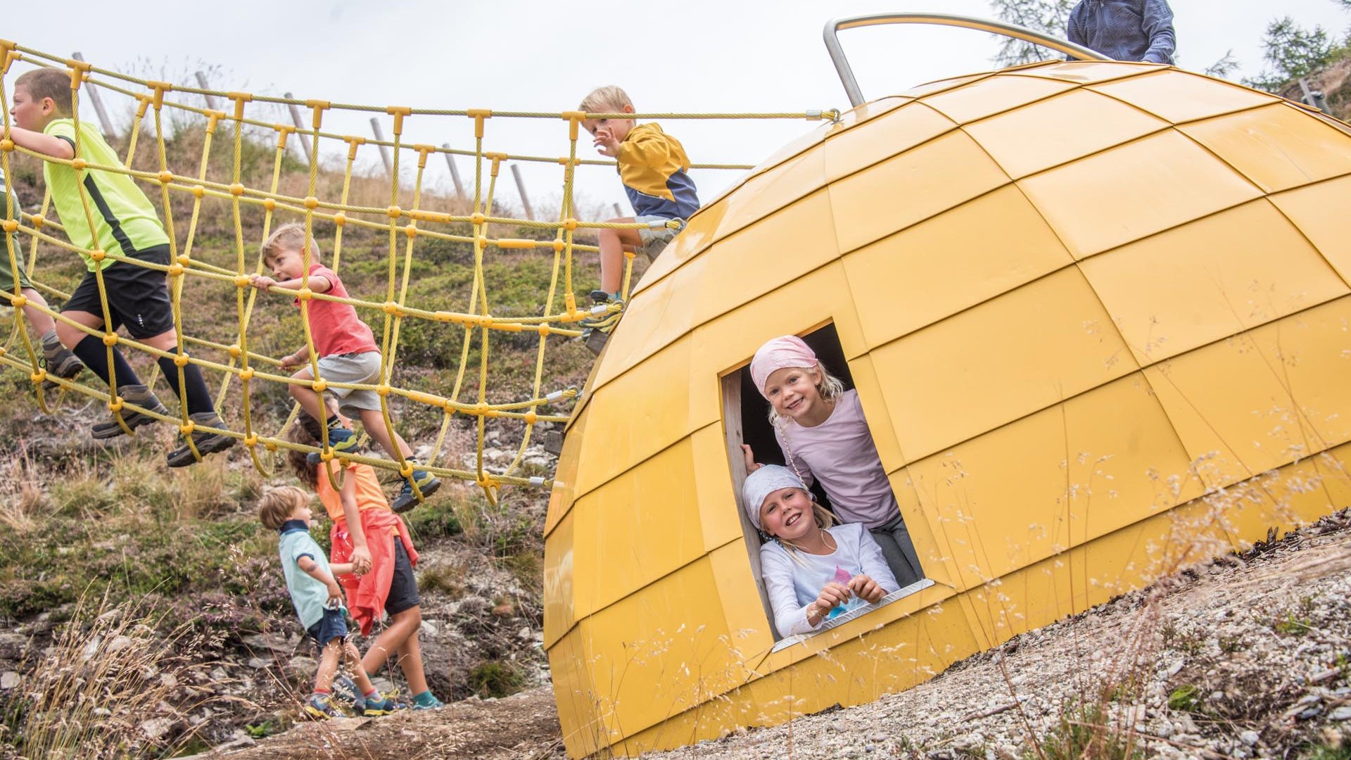 Sonnenpark Gitschberg De foto toont meerdere kinderen die op een speeltuin spelen. Sommige kinderen klimmen over een geel klimnet, terwijl twee meisjes vrolijk uit een geel, bolvormig speelhuisje kijken.
