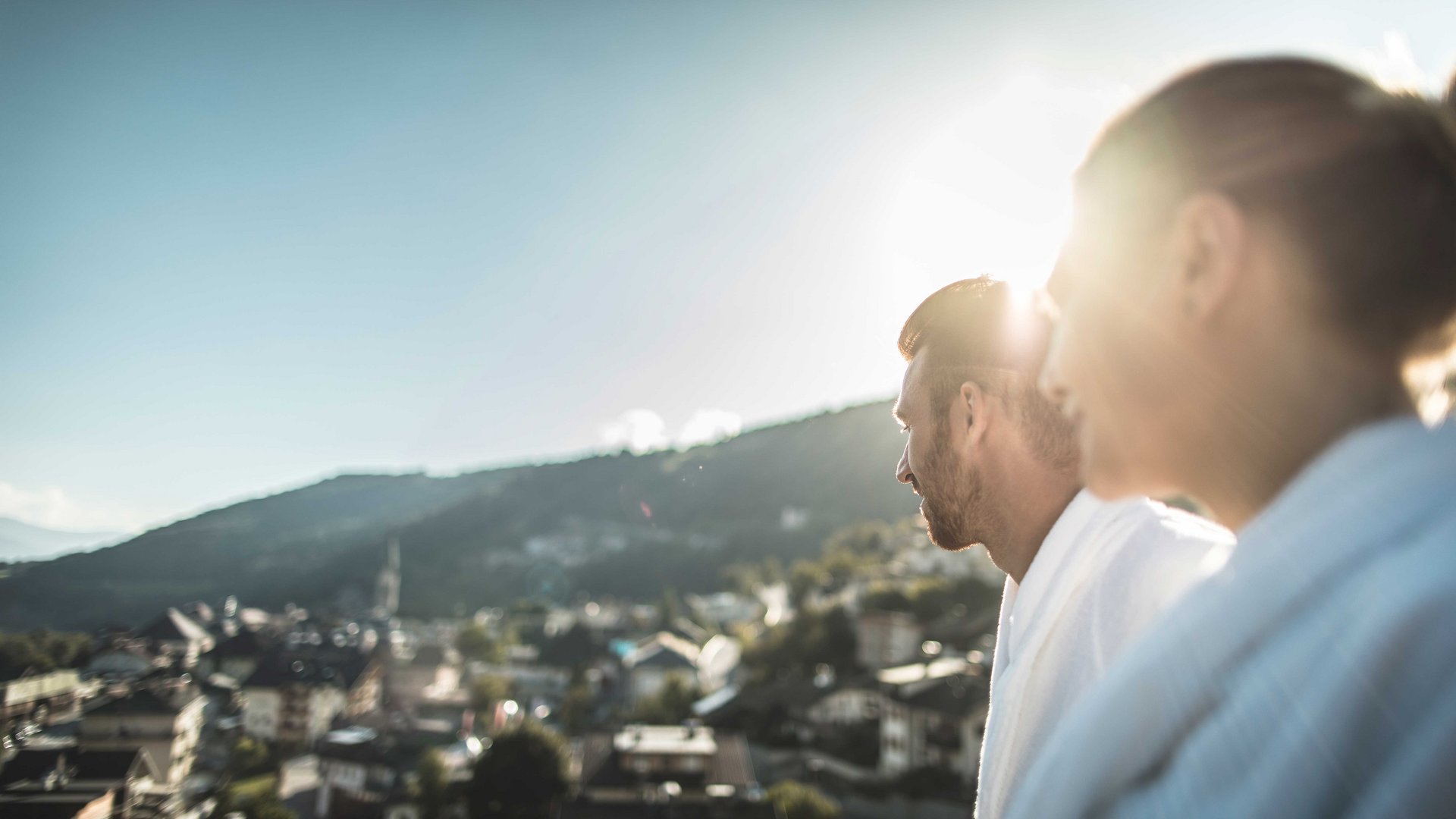 Mühlbach in the Pustertal valley The image shows two people viewed from behind looking into the distance, while the sun shines over a mountain in the background. The people are wearing white bathrobes, and the scene is slightly blurred, creating a soft, dreamy ambiance.