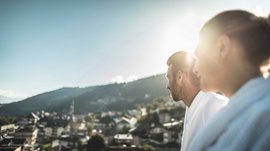 Mühlbach in the Pustertal valley The image shows two people viewed from behind looking into the distance, while the sun shines over a mountain in the background. The people are wearing white bathrobes, and the scene is slightly blurred, creating a soft, dreamy ambiance.