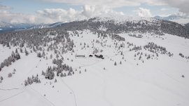 De Rodenecker-Lüsner Alm De afbeelding toont een uitgestrekt, besneeuwd landschap dat wordt doorkruist door glooiende heuvels en kleine bossen. In het midden van de afbeelding bevinden zich enkele berghutten die omgeven zijn door diepe sneeuw, en op de achtergrond rijzen beboste bergen op onder een bewolkte hemel.