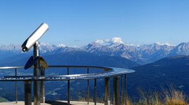 The Gitschberg panoramic platform in Meransen, Italy The picture shows a panoramic viewing platform on a mountain peak, equipped with a permanently installed telescope. In the background, a wide valley stretches out, surrounded by numerous mountain ranges and peaks, under a clear, blue sky.