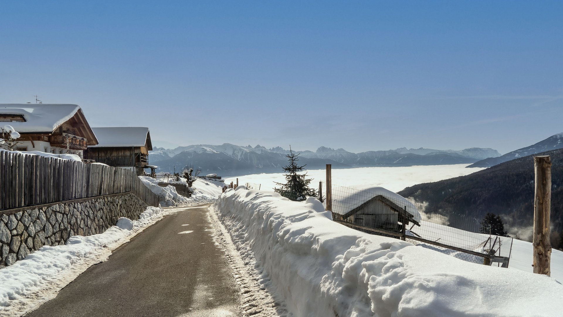 Das Bergdorf Meransen Das Bild zeigt eine schmale, asphaltierte Straße, die durch eine verschneite Landschaft führt. Auf der linken Seite der Straße stehen Holzhäuser und ein Zaun, während im Hintergrund eine dichte Wolkendecke die Täler bedeckt und hohe, schneebedeckte Berge unter einem klaren blauen Himmel zu sehen sind.