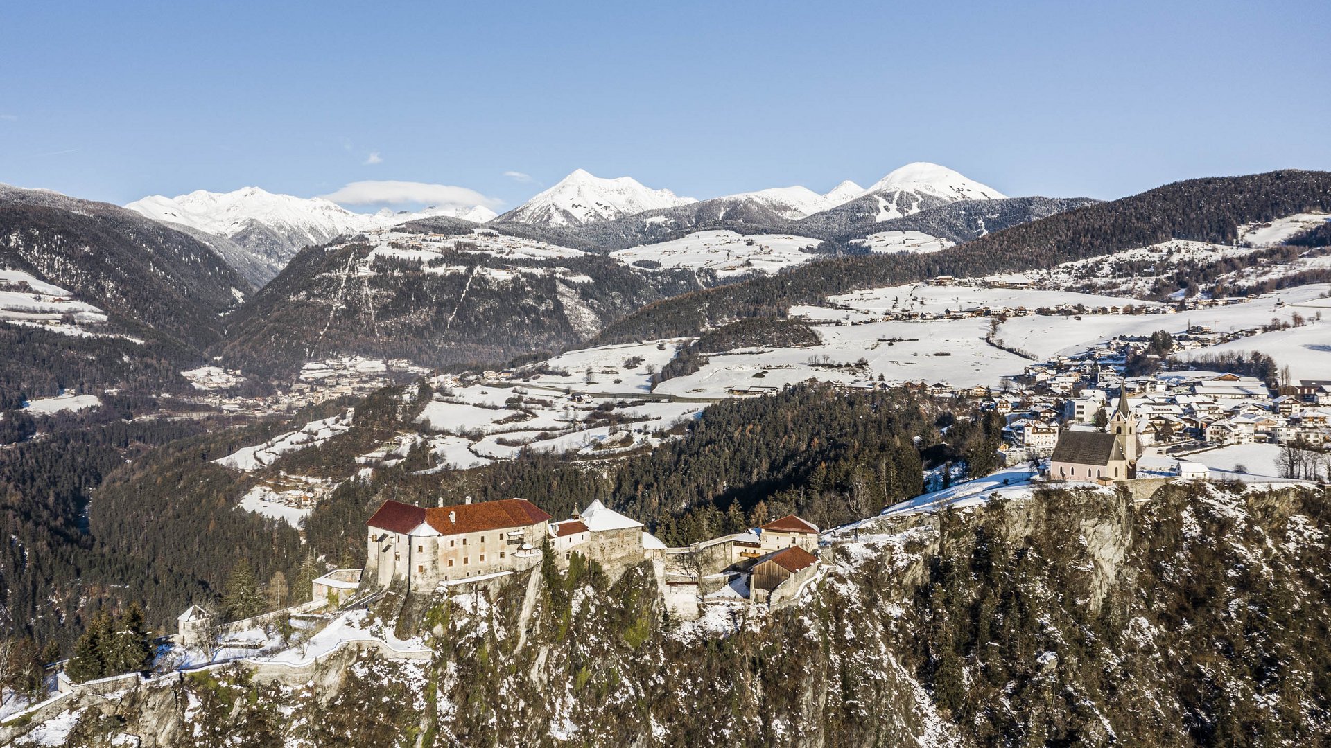 Rodeneck – for a wonderful walking holiday in Italy The picture shows a castle perched on a rugged, snow-covered rock, surrounded by a wintry mountain landscape. In the background, snow-covered mountains stretch under a clear blue sky, while scattered villages and forests can be seen in the valley.